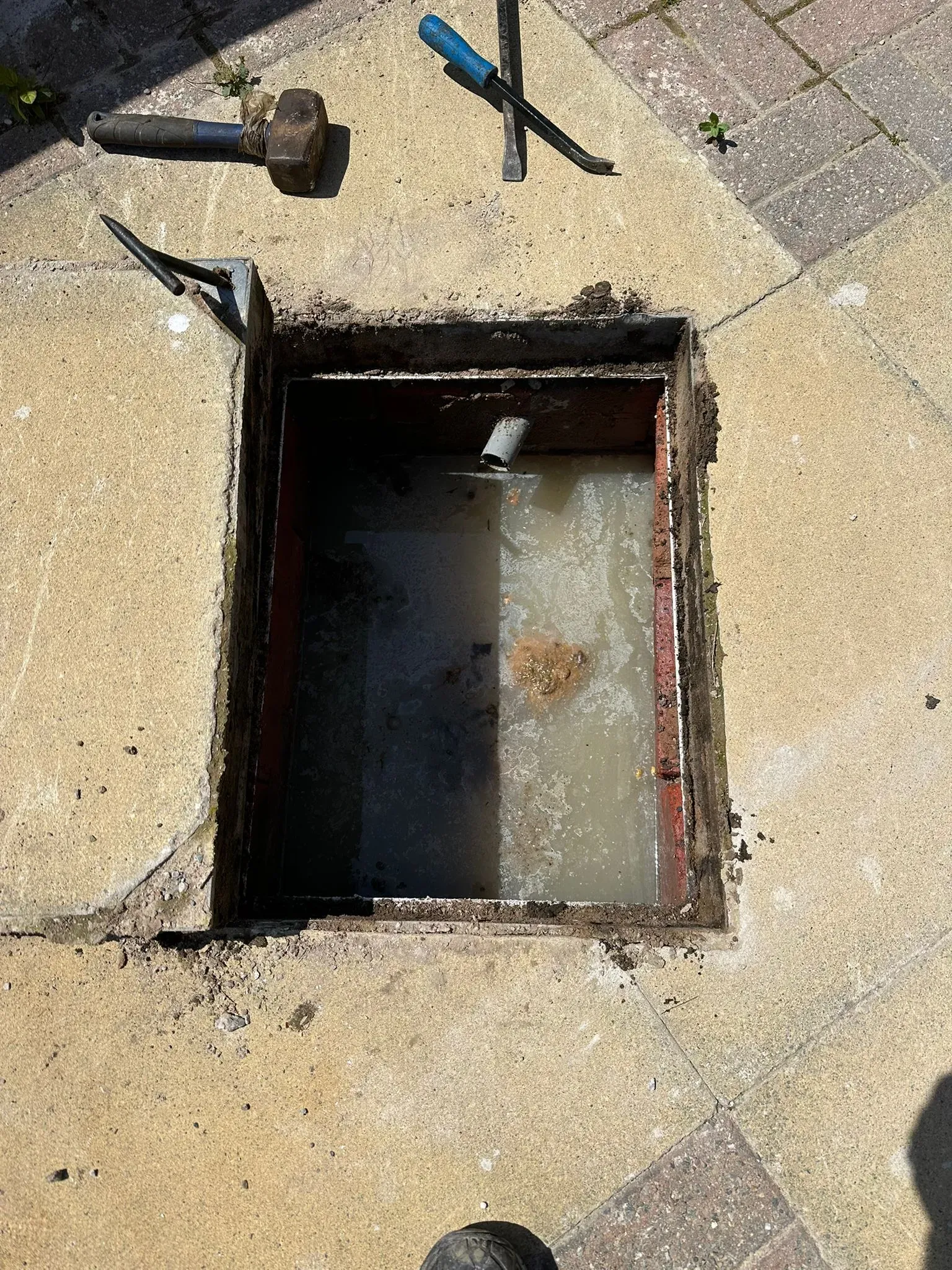 Outdoor drain manhole overflowing with dirty water and drainage tools on the paving, showing a severe blockage.